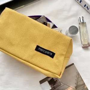 Detailopname van een gele canvas make-uptas met merciparis label en een paarse rits als accent