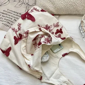 Gros plan sur le sac épaule en toile crème avec des papillons en dentelle brodés rouges.