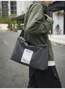 Vista detallada del bolso cruzado gris oscuro con etiqueta llevado por un modelo en calle.