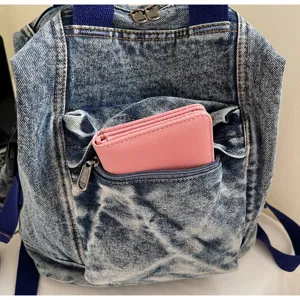 Washed denim backpack detail showing front zipper pocket and pink wallet storage capacity.