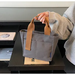 Vista dettagliata della borsa tote in tela grigia, con manici marroni e tasca frontale.