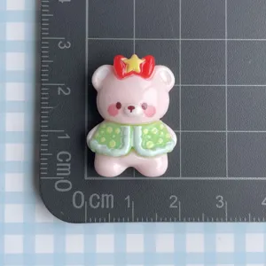 Close-up of cute pink resin bear charm with red bow and green cape for DIY crafting.