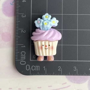 Detail van hars cupcake charm met schattig gezicht en blauwe bloemen op de snijmat.