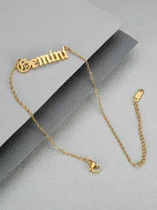 Detailed view of golden Gemini zodiac anklet in Old English font on grey background