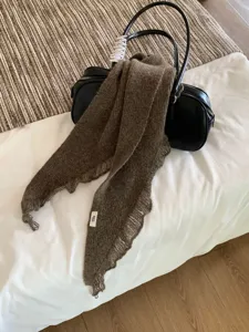 Detail shot of brown knitted frayed scarf styled on black leather bag in cozy bedroom setting.