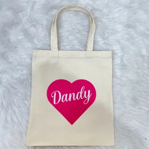 Detailed view of a cream canvas tote bag featuring a pink heart and Dandy script print