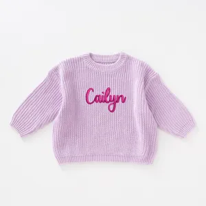 Macro detail of the soft purple knit texture and pink name embroidery on the baby sweater.