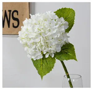 Close-up detail of white artificial hydrangea flower with green leaves in glass vase.