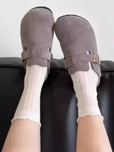 Detail view of breathable white lace-trim socks worn with casual grey suede mule shoes outdoors.