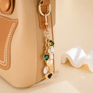 Detail view of heart-shaped charm with green gems and pearls on a tan leather bag