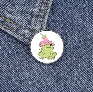 Cute cartoon frog metal brooch with a pink flower hat and peace sign design. Detail shot.