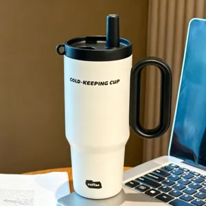Close-up detail of white insulated tumbler with black handle and straw lid on wooden desk.