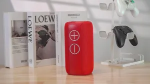 Portable red cylinder bluetooth speaker with fabric mesh and volume buttons for outdoor use.
