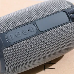 Altavoz Bluetooth inalambrico portatil con tejido de malla gris y botones de control de goma.