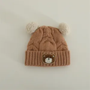 Infant knit beanie detail, brown cable knit texture with cute bear patch and fluffy pom poms.