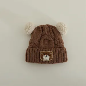Detailed view of brown baby knitted hat with cute white pom-poms and lovely bear patch.