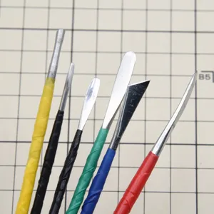 Close-up detail of stainless steel clay sculpting tool heads with colorful handles on grid mat.