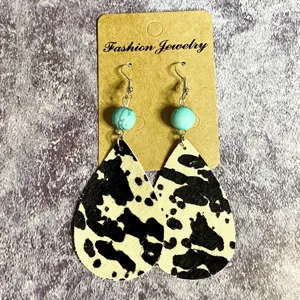 Gros plan de boucles d'oreilles pendantes en cuir imprimé vaches avec perle de turquoise.