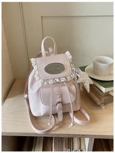 Detail shot of a pink leather backpack with white lace trim and metal plaque on a wooden table.