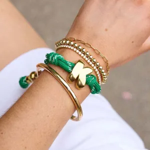 Detailed close-up shot of stacked gold chain bracelets featuring a green rope puffy letter K charm.