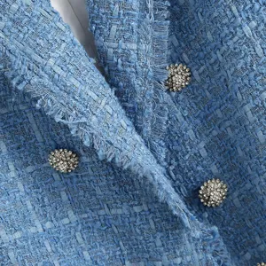 Close up view of light blue tweed blazer fabric, fringe trim and sparkling rhinestone buttons.