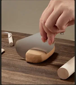 Stainless steel clay scraper tool cutting pottery clay block on wooden workspace.