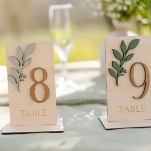 Beaux numéros de table en bois avec feuilles vert sauge pour décoration mariage détails.