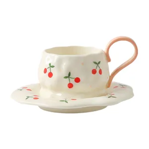 Handmade ceramic coffee cup and saucer set with cute red cherry pattern and pink handle.