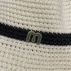 Detailed view of white woven straw sun hat with black ribbon and sparkly rhinestone letter m.