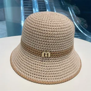 Beige straw bucket hat with contrast brown stripe and metal letter detail for summer sun protection.