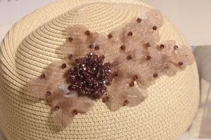 Detailansicht eines schicken beige Strohberets mit eleganter Perlenblume und Strasssteinen.