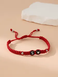 Dettaglio di bracciale intrecciato rosso con perla lettera 'S' nera e icona a cuore.