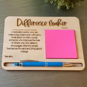 Wooden sticky note holder with inspirational quote text, pink memo pad and blue pen desk decor.