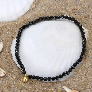 Close-up detail of faceted black spinel anklet with small golden ball charm on a white seashell