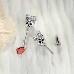 Detailed view of silver skull ear studs with bows and a faceted red teardrop gemstone pendant