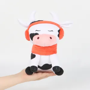 Suave peluche de vaca de dibujos animados con orejeras y bufanda naranjas en la mano