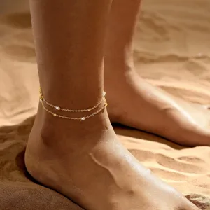Detailansicht einer doppellagigen goldenen Fußkette mit Perlen auf sonnigem Sand