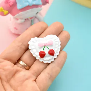 Close-up of white heart resin accessory with pink bow and red cherry DIY craft detail