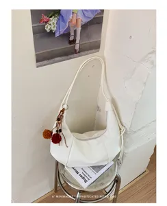 Stylish white croissant-style shoulder bag with decorative pom-pom charms in a minimalist setting