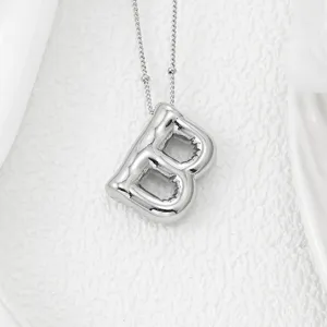Gros plan détail d'un pendentif collier lettre B argentée rebondie sur un tissu blanc.