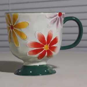 Keramiktasse mit erhabenem Blumenmuster, hohem Fuß und grünem Henkel zum Frühstück.