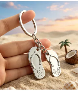 Detailed macro shot of a silver metal flip-flop keychain held against a sandy beach sunset.