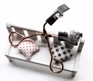 Detail view of rose gold magnetic eyelash curler and soft false lashes set on miniature bench.