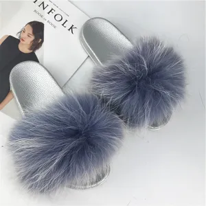 Detail view of stylish silver PVC slides featuring large fluffy grey raccoon fur pom-poms