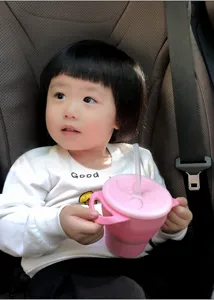 Adorable toddler using pink silicone snack cup with straw and handles in car seat detail.