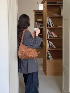Detailfoto van bruine geplooide schoudertas met strikdesign in een gezellige bibliotheekomgeving.