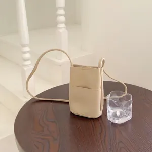 Close-up of minimalist beige leather crossbody bag on dark wooden table with clear glass cup.