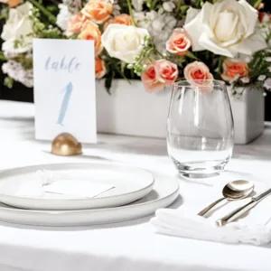 Detalle de mesa elegante con platos blancos de cerámica, cristalería y decoración floral.