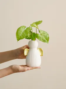Close-up detail view of white ceramic angel vase with gold wings holding fresh green plant.