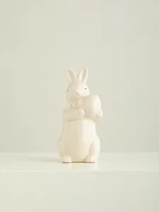 Close-up of white ceramic bunny figurine, cute rabbit holding egg decor for desktop.
